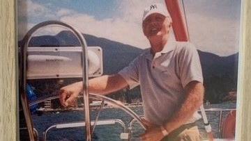 An old photo of a man in a white yankees cap with mountains behind him standing on a boat on a sunny day, smiling. 