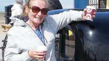 A woman in her late 60s smiles at the camera while leaning against a black cab. She is holding two hot drinks in disposable paper cups in each hand, has light ginger hair tied back and is wearing a grey zip-up hoody and sunglasses.