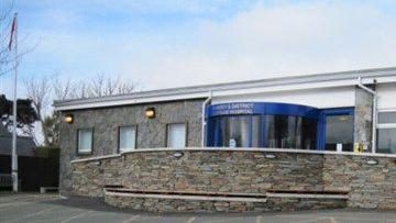 The front entrance to the hospital, which is a one-storey building with a blue revolving door. There is a stone wall in front of it separating it from the car park.