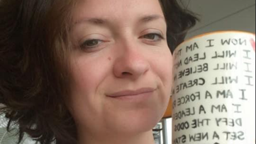 Woman with shoulder-length brown hair and a grey top with a mug in her hand.