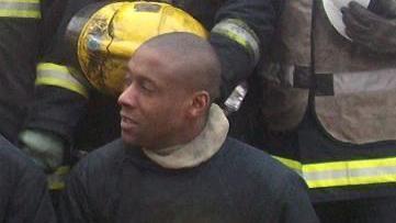 Jez Daniels in his firefighter's uniform, holding a yellow helmet and with colleagues behind him