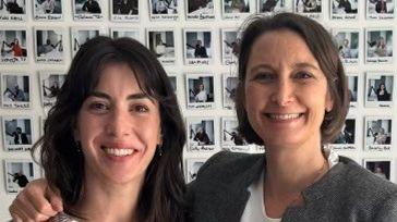 Two women with brown hair smile at the camera. The woman on the right has her arm around the woman on the left. Dozens of Polaroid photos are stuck on the wall behind them.