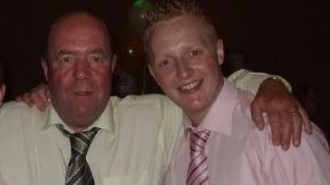Colin Bell, a man with a shaved head, with his arm around his adult son Kevin, who had short ginger hair. Colin is wearing a yellow shirt with a striped green/grey tie and Kevin is wearing a pink shirt with a pink and red striped tie.