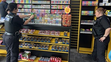 Two female police officers stand in a shop which has yellow and black shelves lined with products