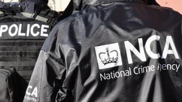 NCA officer dressed in black with agency's logo on jacket, alongside police officer dressed in black 