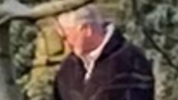 A grainy CCTV clip of a man with white hair, wearing a dark coat. He is pictured behind a tree branch.