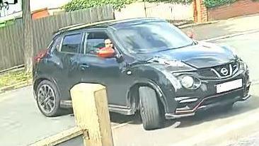 A black Nissan Juke with red wing mirrors drives towards the camera. There is a man in the driver's seat with the window partially down. 