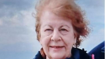 Close-up of Mary White smiling at the camera. She has red hair, which is now grey, and a blue jacket. The sky fills the background.