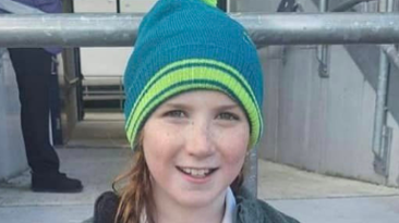 A young girl sits and smiles at the camera. She is wearing a green and blue beanie hat, a green coat and rugby shirt.