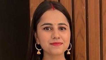 Harshita Brella is standing outside against a wooden wall. She is looking at the camera and is smiling.