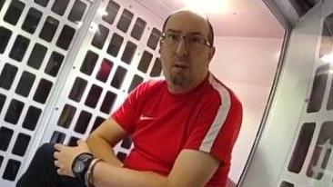 Adam Evans is sitting in the back of a police van. He is handcuffed and wearing glasses and a red Nike top.
