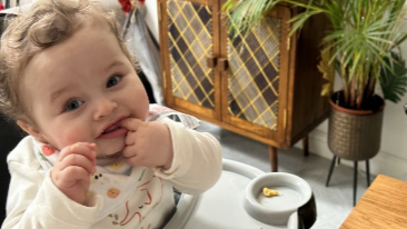 Baby Preston Davey has a finger in his mouth as he is eating in a high chair. He has brown curly hair and he is wearing a cream baby grow with an elephant on the front and a bib.