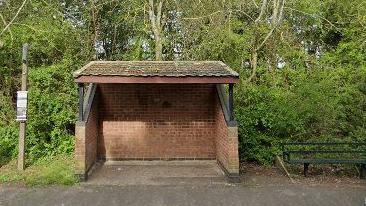 A red-brick empty bus shelter, with a bench to its left and bus sign to its right. 