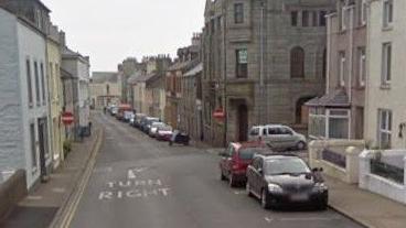 A view looking down High Street in Port St Mary against the flow of traffic. It is a narrow street with tall buildings on either side and cars parked on the right.