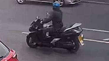 A person wearing a blue helmet and dark clothing rides a black moped along a road, with cars visible nearby.
