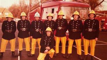 A group of eight male firefighters standing in front of a bright red fire truck. Seven of the firefighters are stood in a row, with one of the firefighters crouched on one knee in the middle, in front of those standing. All of the firefighters are wearing black buttoned jackets with big, brown belts around their torsos, and are also wearing bright yellow trousers and brown boots. They are in a car park, with the fire truck positioned in the background to their right, front-on.