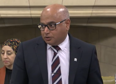 Bald man of Asian heritage with black glasses wearing white shirt dark blazer and striped tie