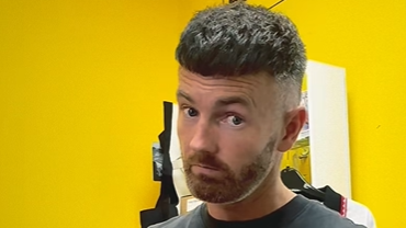 Chris has hair shaved close at the sides and longer on top. He is wearing a black vest and has his index finger raised towards the camera. Behind him is a yellow wall.