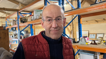 Rob Byass. A 69-year-old man with glasses wearing a blue turtle neck and red puffer gilet. He is standing in a barn. To his left is a shelf holding several donated clocks. 