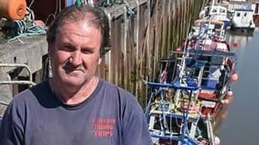 John Worthington with brown hair wearing a navy blue t-shirt standing above Jubilee Quay with fishing boats in the water. 