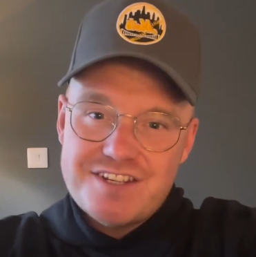 A selfie of Ryan Forey. He is wearing a grey cap with a logo with a cityscape on it and a black top, and has rounded glasses.
