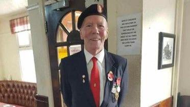 An elderly man wearing a dark jacket and red tie and Army beret. His lapel has a poppy and medals on and he is smiling at the camera. 