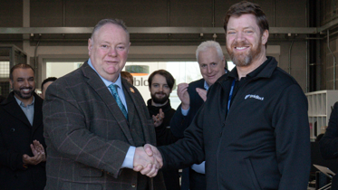 Two men, one in a grey checked suit, one in a branded fleece, shake hands in front of a group of people in a workplace.