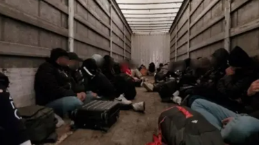 Dozens of people are seated on the floor inside a large lorry. Their faces are blurred in the image and many appear to be male in jeans and hooded tops and jackets.
