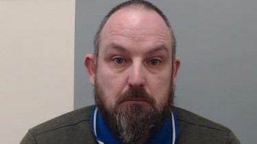 A man with a bald head and grey beard and dark eyes. He has a blue collar on and grey top. He is looking at the camera and behind him is a white and blue wall. 