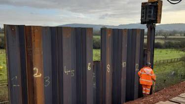 A National Rail worker in a bright orange hi vis jacket and trousers installs piling: tall, vertical pieces of metal with spray painted numbers on each piece - to help stabilise a road surrounded by fields.