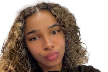 A selfie of a young woman with curly hair in front of a white backdrop.