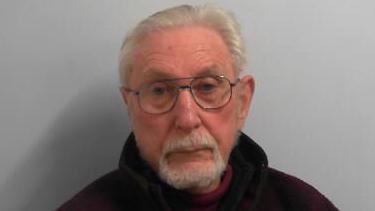 Mugshot of David Hampshire against a grey background. He is elderly with wire frame glasses, a white goatee and white combed-back hair. He is wearing a black hoodie.