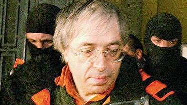 A picture of Gregorian Bivolaru - wearing a green jumper and an orange and black coat, with long, thin white hair - being arrested in Romania in 2004