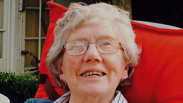 A woman with short grey hair and glasses, smiles while looking up. She is sitting in a chair outside, with a red seat cover behind her head