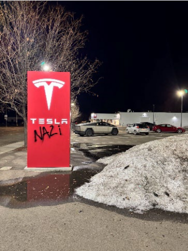 A Tesla dealership with the word "Nazi" spray painted on the sign. Cars and snow banks are visible in the photo.