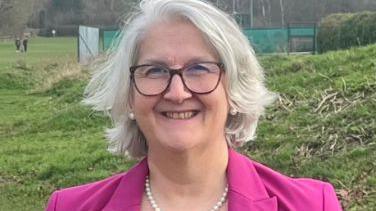 Head and shoulders photo of Edna Murphy in a park with grass fields around her. She wears a pink blazer, white pearl necklace, matching earrings and glasses. She has grey hair which reaches just above her shoulders and is smiling at the camera. 