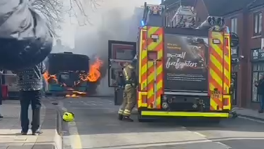 A fire engine is stopped in the middle of a street, with a bus nearby which has flames emerging from it engine compartment. There are a number of people on the pavements either side of the street who are filming or photographing the scene on their phones.