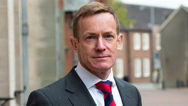 Alan White has short light brown hair. He is posing in a street and his head and shoulders are visible. He is wearing a black suit jacket with a white shirt and blue and navy striped tie