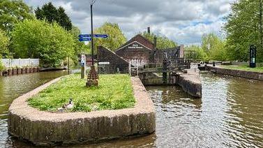 Part of a canal with a lock and building in the background. A central island has a pillar on it with three blue signs.