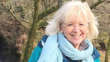 Catherine Sams is standing in a woodland on a sunny day. She is wearing clear-framed glasses, pearl earrings, a blue scarf and a blue raincoat. 