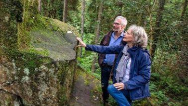 Older man and women, both with grey hair, gaze at a two metre high rock shaped like a chair. The woman is rested the fingertips of her outstretched arm on the slippery, mossy stone.