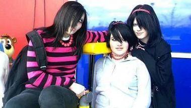 A picture of three girls all with straight black hair and a fringe covering half their faces. The girl on the left wears a pink and black striped top and black trousers while sitting on a children's ride. A black rucksack falls off her shoulder.
The girl in the middle has a grey hoodie and a red beaded necklace. Behind her to the right a girl wears a black hoodie, she has long black hair and a pink headband. 