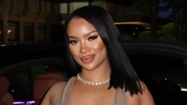 A woman in a light brown dress stands in front of a car, with a shell necklace and straight black hair.