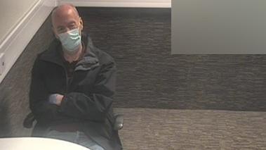 An older man sits with his arms crossed and wearing a face mask in a carpeted room in a police station. 