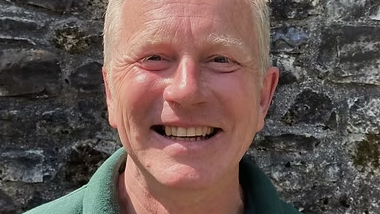 Andy smiles into the camera. He is standing in front of a grey stone wall. He has blonde hair and a green shirt. 