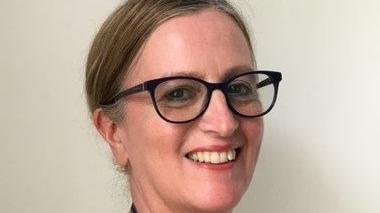 A woman with blonde hair smiles at the camera. Her hair is tied back and she is wearing dark-coloured oval glasses.

