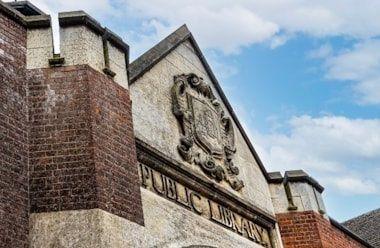 Red brick and stone library building, showing close-up of engraved "public library" sign above the door, with a cote of arms. Also visible is a crenelated roof design 