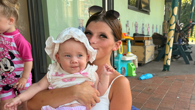 A mother wearing a white top is smiling for a photo while holding her baby girl. Another little girl can be seen near them. It is sunny.