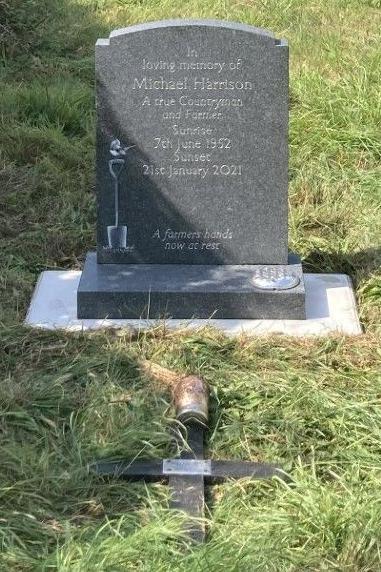 The whole of the grey marble memorial stone can be seen with the wooden cross lying face down on the grass in front of it. The stone is carved with: In loving memory of Michael Harrison, a true countryman and farmer, sunrise 7th June 1952, sunset 21st January 2021, a farmer's hands now at rest. There is a a carving on the left which shows a spade with a bird resting on its handle.