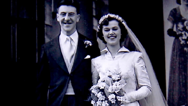 A black and white photo of a bride and groom. The man is waring a black morning suit and the woman is wearing a white collared dress and veil while holding a bouquet of flowers
